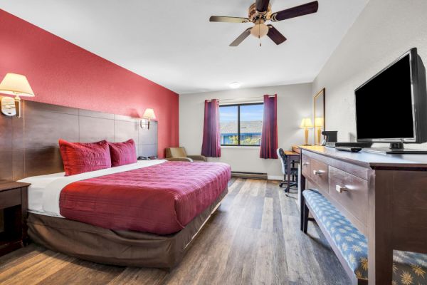 A hotel room with a king-size bed, red accents, TV, desk, and window with a view. There's a chair and a ceiling fan.