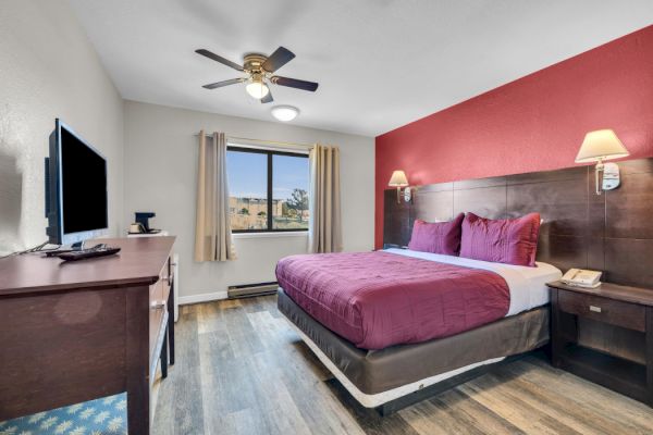 A cozy hotel room with a double bed, red accents, wall lamps, TV, window, and wooden floor, creating a welcoming atmosphere.