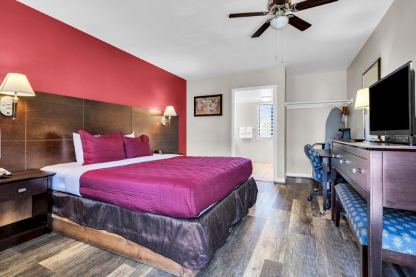 The image shows a hotel room with a bed, TV, desk, chair, ceiling fan, and a red accent wall.
