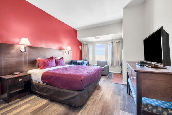 A hotel room with a red accent wall, bed, TV, and seating area by the window, combining modern decor with comfort.