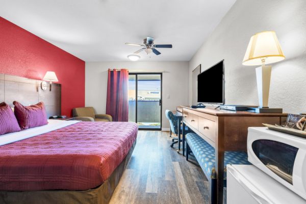 A hotel room featuring a large bed, TV, desk, microwave, and a view through sliding glass doors, with a red accent wall and modern decor.