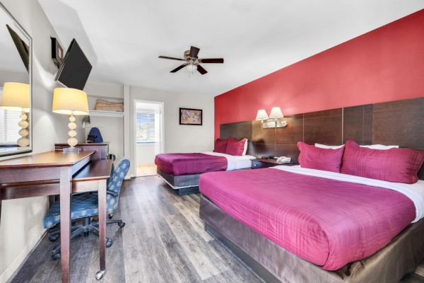 The image shows a hotel room with two double beds, a desk, chair, wall-mounted TV, lamps, and a ceiling fan against a red accent wall.