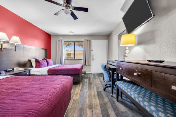 The image shows a hotel room with two beds, a desk, a wall-mounted TV, a lamp, and a window with curtains.