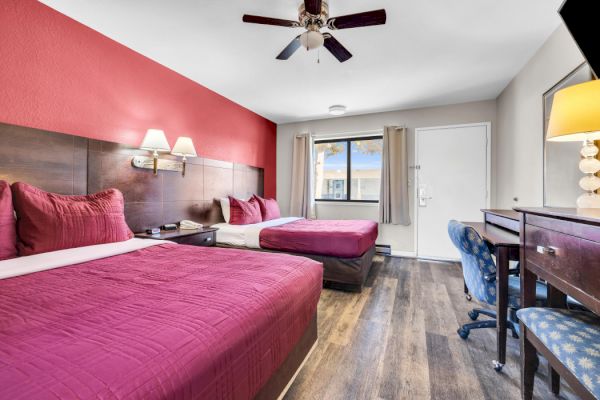 A hotel room with two beds, red accents, a desk, chair, lamp, and ceiling fan, with a window and door visible.
