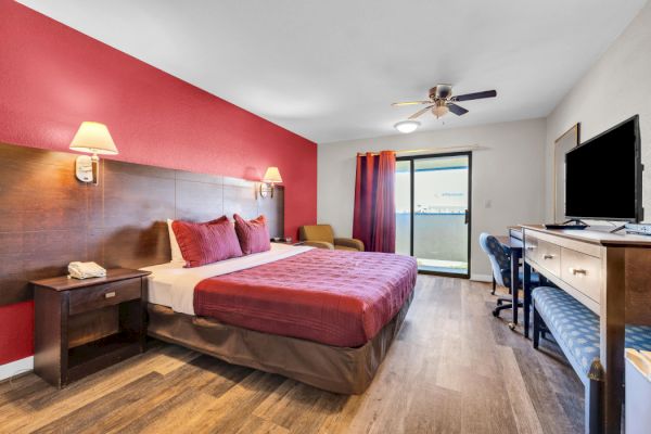 The image shows a hotel room with a bed, TV, desk, chair, and window with a view, featuring red and brown decor.