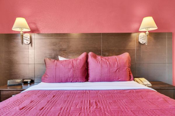 A bed with pink bedding, two pillows, and wall lamps in a room with a pink wall, along with a phone and a clock on the bedside table.