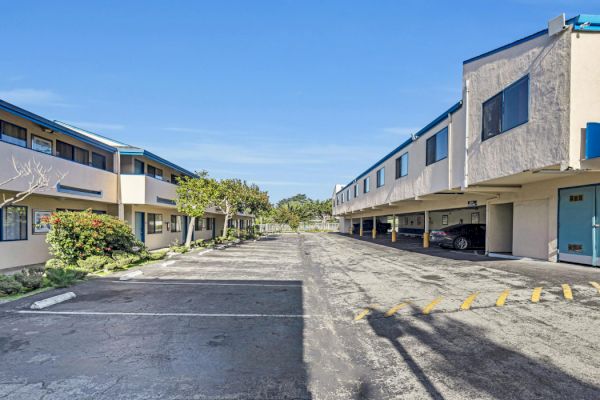 The image shows a paved driveway between two apartment buildings with covered parking on the right and some greenery on the left.