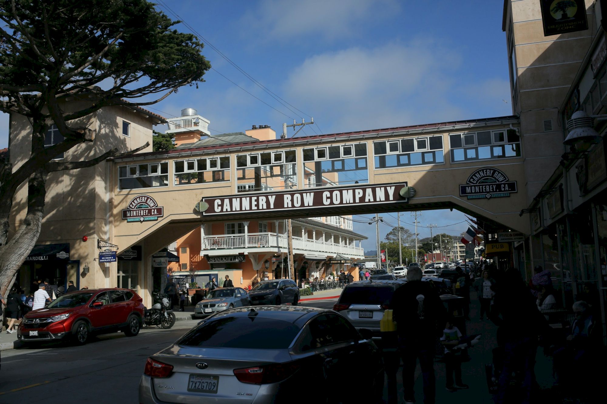 Busy street scene at Cannery Row, with cars, pedestrians, and the Cannery Row Company bridge under a partly cloudy sky.