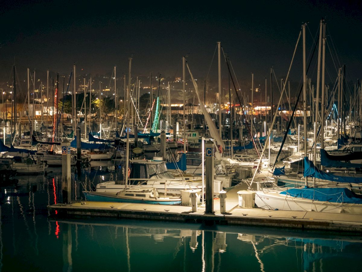 The image shows a marina at night with numerous sailboats docked and city lights in the background reflecting on the water.
